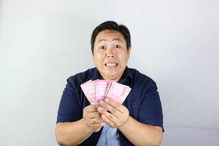 Shocked Indonesian man in blue shirt holding stacks of money with mouth wide open, wow expression