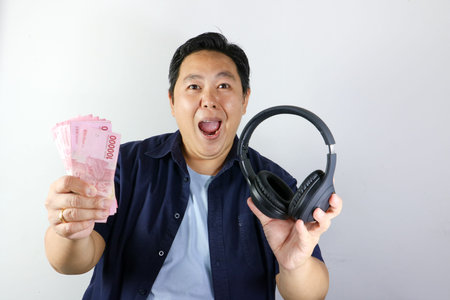 Wow expression of Asian man holding black headphones and a stack of Indonesian Rupiah cash against a...