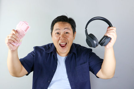 Wow expression of Asian man holding black headphones and a stack of Indonesian Rupiah cash against a white backgroundの写真素材