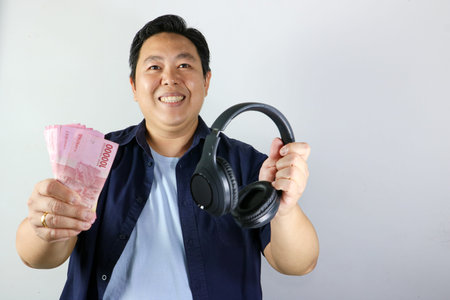 Smiling Asian man holding black headphones and a stack of Indonesian Rupiah cash against a white backgroundの写真素材