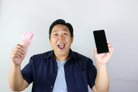 Portrait of surprised asian man holding a smartphone and cash with wow expression
