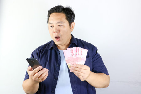 Curious Asian man holding a smartphone and a stack of Indonesian Rupiah cash against a white...
