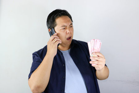 Curious Asian man holding a smartphone and a stack of Indonesian Rupiah cash against a white...