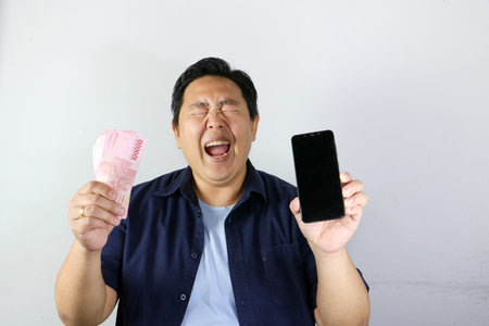 Portrait of amazed Indonesian man reacting wow to a huge win holding mobile phone and a stack of...