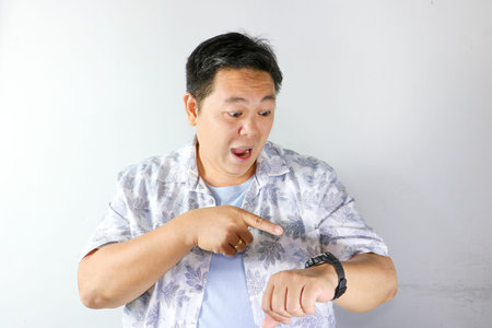 Surprised Asian man looking at his wrist watch and pointing at it with open mouth on white...