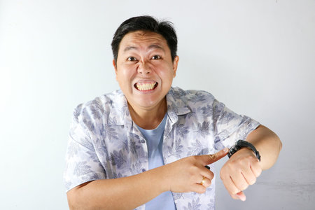 Shocked Asian man looking at his watch and pointing at it on a white backgroundの写真素材