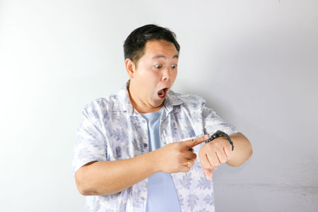 Surprised Asian man looking at his wrist watch and pointing at it with open mouth on white...