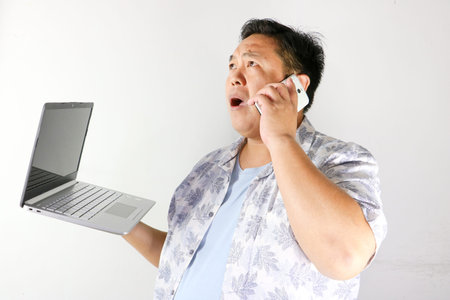 Portrait of Asian man with a curious expression talking on a phone while holding a silver laptopの写真素材