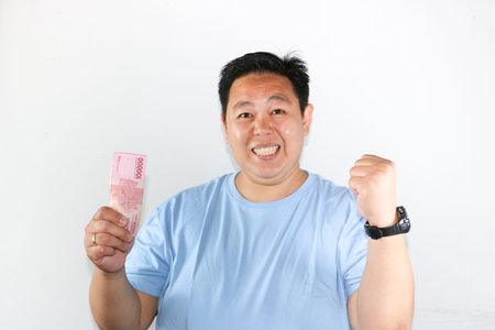 Shocked Asian man holding Indonesian Rupiah banknotes and making a winning fist gesture with...