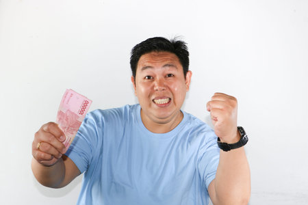 Shocked Asian man holding Indonesian Rupiah banknotes and making a winning fist gesture with...