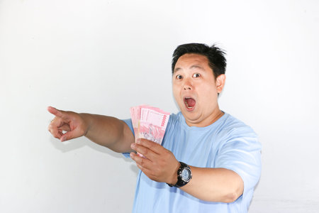 Shocked Asian man holding piles of Indonesian Rupiah banknotes toward the camera with wide open...