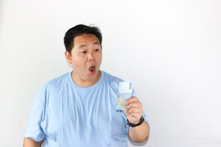 Shocked Asian man holding piles of Indonesian Rupiah banknotes toward the camera with wide open...