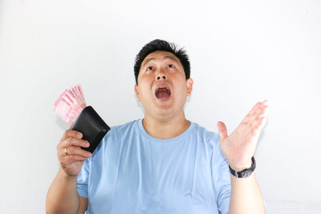 Shocked Asian man holding piles of Indonesian Rupiah banknotes toward the camera with wide open...
