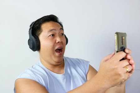 Cheerful asian man with mouth open in surprise watching exciting media content on a mobile phoneの写真素材