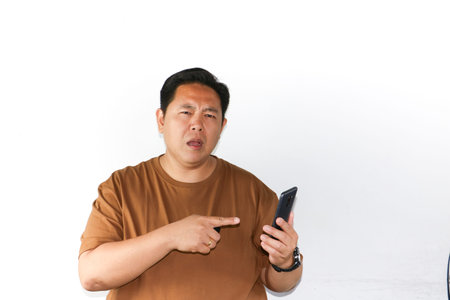 Puzzled Asian man looking at a smartphone with blank screen and thinking gesture on white backgroundの写真素材