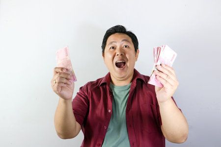 Happy Asian man holding cash of Indonesian Rupiah banknotes smiling at camera on white backgroundの写真素材