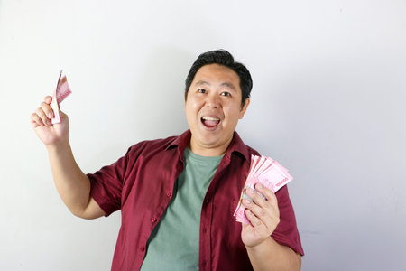 Excited asian man in red shirt shouting for joy while holding stacks of indonesian rupiah banknotesの写真素材