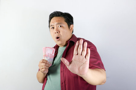 Shocked asian man in red shirt holding indonesian rupiah and making a stop gesture with his handの写真素材