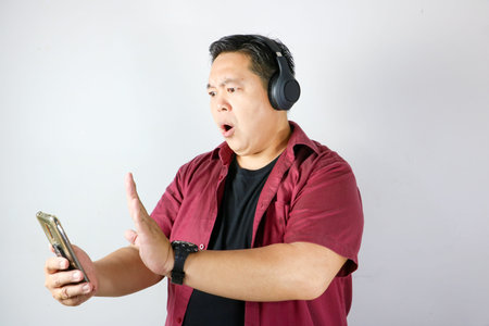 Shocked Indonesian man wearing headset and holding smartphone showing rejection sign with his handの写真素材