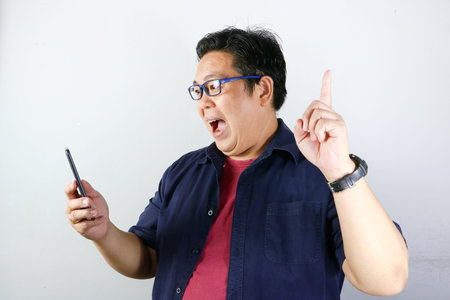 Excited Asian man in blue shirt gets a brilliant idea while looking at his smartphone on white...