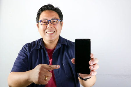 Smiling handsome Asian man in blue shirt points finger at black smartphone screen