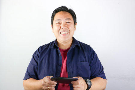 Smiling handsome Asian man in blue shirt points finger at black smartphone screenの写真素材