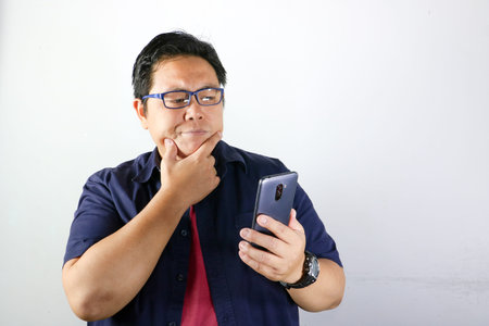 Portrait of Curious Asian man in blue shirt touching his chin while looking at a smartphone on white...