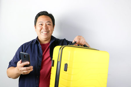 Portrait of a smiling Asian man on traveling holding a yellow suitcase and showing a smartphone