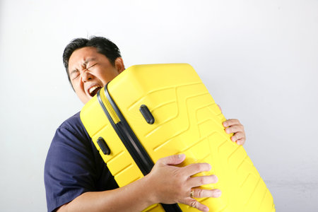 Portrait of a struggling man carrying a heavy yellow suitcase on his shoulder against a white...