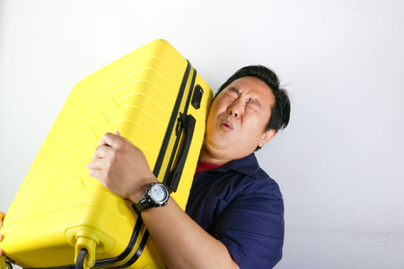 Portrait of a struggling Asian man carrying a heavy yellow suitcase on his shoulder against a white backgroundの写真素材