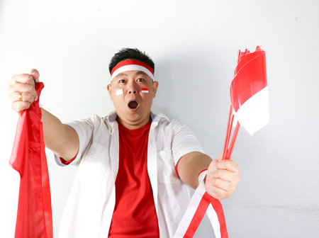 Enthusiastic Indonesian man holding small national flag and showing winning gesture on white backgroundの写真素材