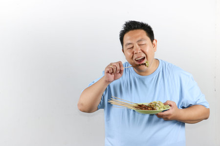 Portrait of Indonesian man eating delicious chicken satay with peanut sauce and lontong rice cakesの写真素材