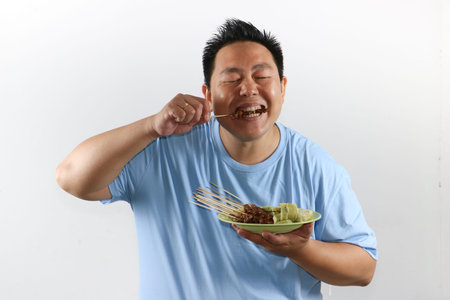 Portrait of Indonesian man eating delicious chicken satay with peanut sauce and lontong rice cakesの写真素材