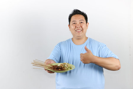 Happy Asian man holding a plate of authentic Indonesian chicken satay with peanut sauce and lontong...