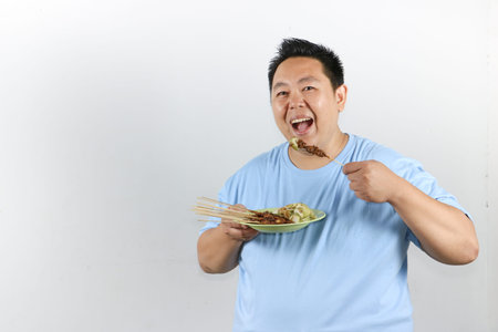 Happy Indonesian man eating authentic chicken satay with peanut sauce and lontong, isolated on white backgroundの写真素材