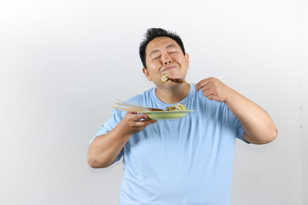 Happy Indonesian man eating authentic chicken satay with peanut sauce and lontong, isolated on white backgroundの写真素材