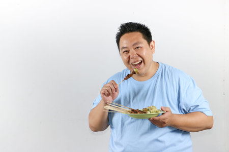 Portrait of an Asian man enjoying delicious grilled chicken skewers or sate ayam, isolated on white background.の写真素材