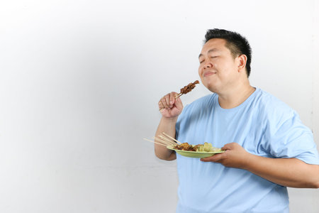 Happy Indonesian man eating authentic chicken satay with peanut sauce and lontong, isolated on white backgroundの写真素材