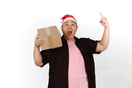 Cheerful Asian man wearing Santa hat holding a cardboard box package on white background, holiday...