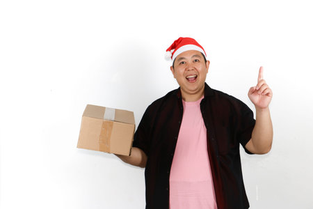 Excited Asian man wearing santa hat holding a cardboard box package on a clean white background