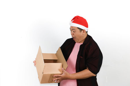 Excited Asian man wearing santa hat holding a cardboard box package on a clean white background