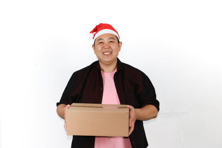 Excited Asian man wearing santa hat holding a cardboard box package on a clean white background