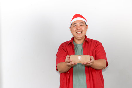 Portrait of happy Asian man wearing a red Santa hat holds a brown cardboard gift box with both...