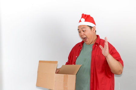 Portrait of happy Asian man wearing a red Santa hat holds a brown cardboard gift box with both...