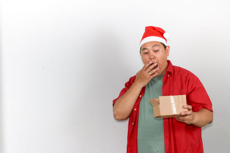 Portrait of happy Asian man wearing a red Santa hat holds a brown cardboard gift box with both hands.の写真素材