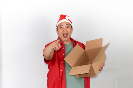 Portrait of happy Asian man wearing a red Santa hat holds a brown cardboard gift box with both...