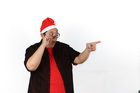 Portrait of enthusiastic Asian man wearing Santa hat pointing to empty space isolated on white backgroundの写真素材