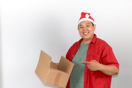 Portrait of happy Asian man wearing a red Santa hat holds a brown cardboard gift box with both...