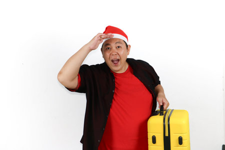 Portrait of enthusiastic Asian man wearing Santa hat and holding yellow luggage on white background studioの写真素材