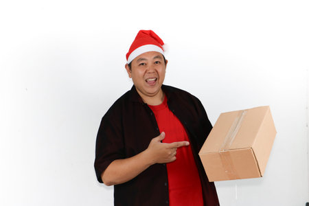 Portrait of cheerful Asian man in Santa hat holding cellphone and pointing to blank screen on white...